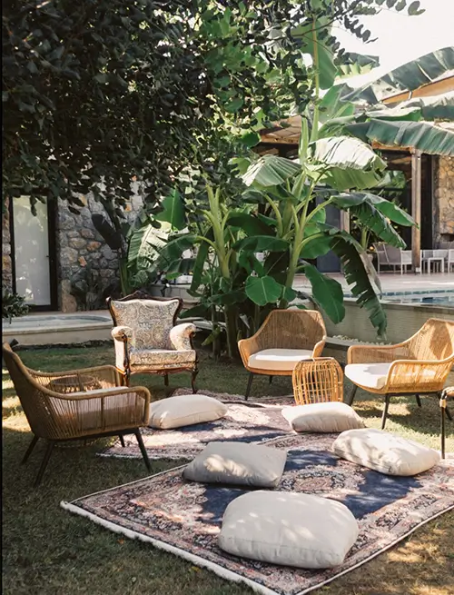 Wicker chairs on a chic garden rug with cushions under a pergola, stone villa exterior, and tropical plants.